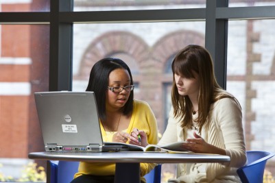 Two women seated at a table with a laptop, reading from a magazine.